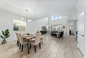 A spacious living area with a dining table, seating, and a large window. - Home & Storey Lake Water 5br/5ba - Park Amenities (Kissimmee)