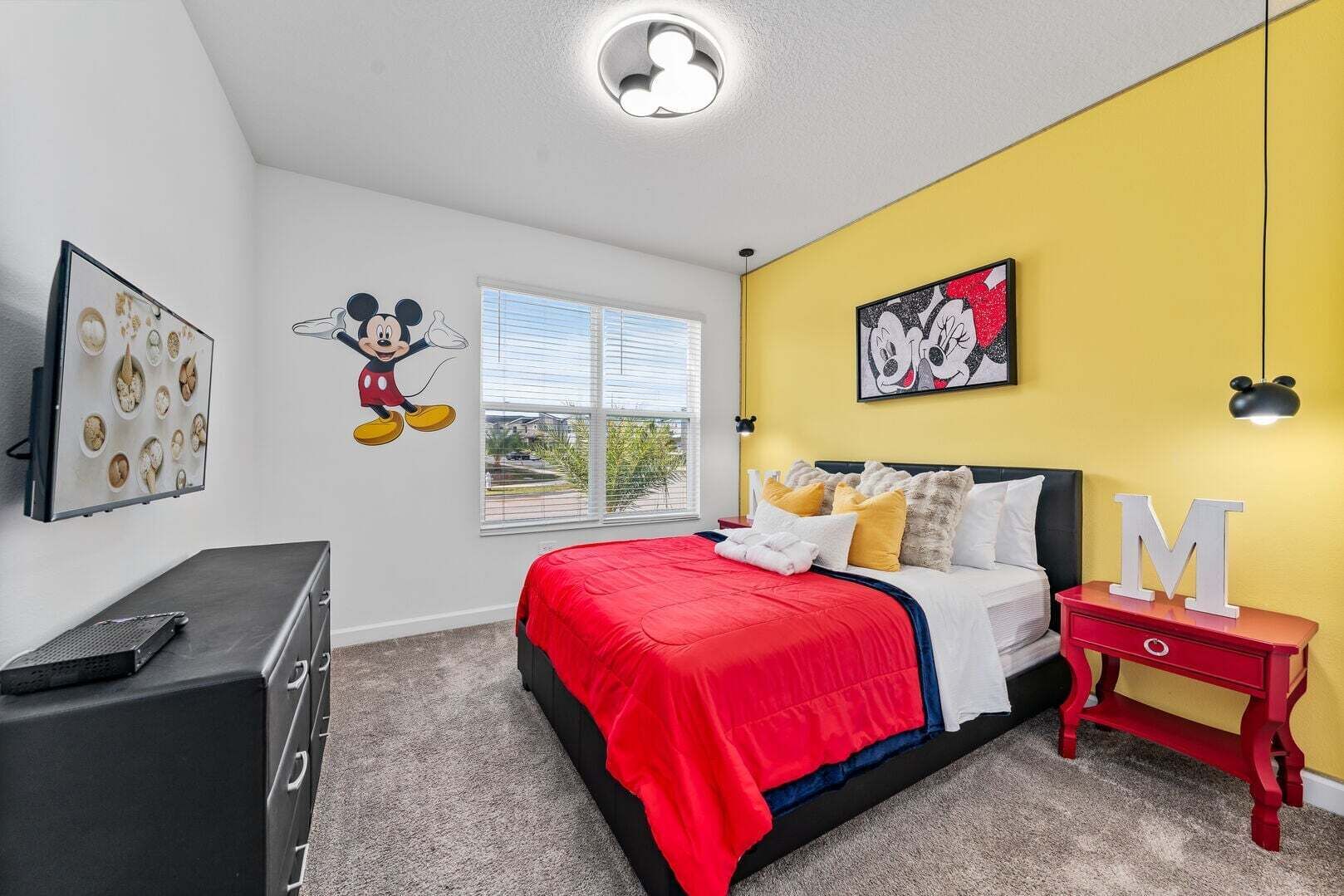 A bedroom with a bed, a nightstand, a dresser, Mickey Mouse wall decal, a window, and a yellow accent wall.