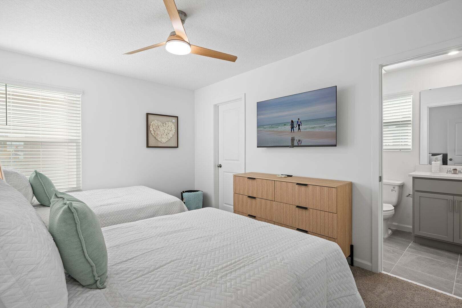 A bedroom with two beds, a wooden dresser, a ceiling fan, a framed picture, and a bathroom with a toilet and sink.