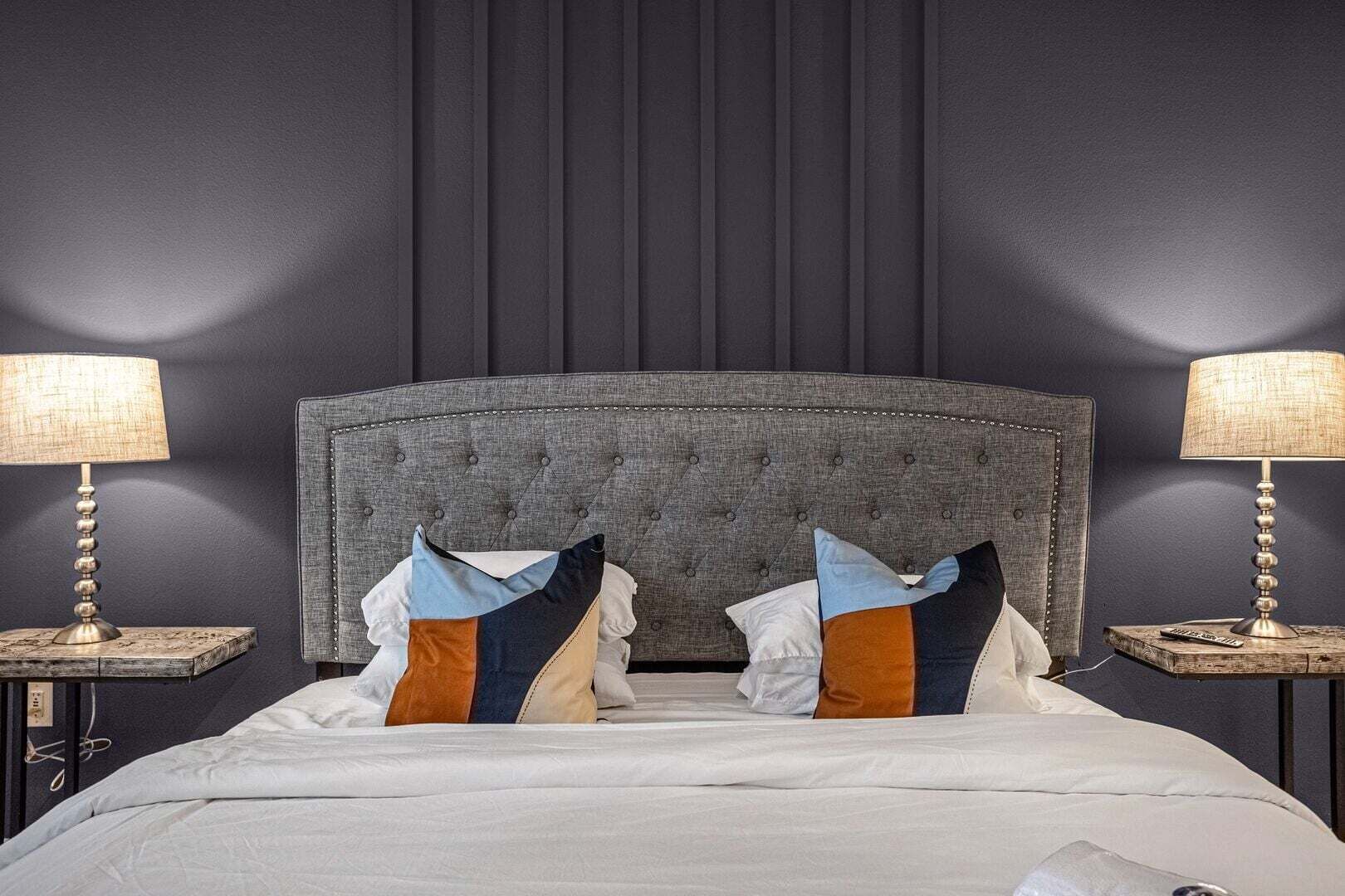 Habitación con cabecera capitonada, dos mesitas de noche con lámparas y una cama con varios cojines.