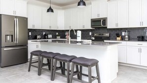 A modern kitchen with white cabinets, stainless steel appliances, a large island with bar stools, and pendant lighting. - Home Away From Home Private Pool (Davenport)