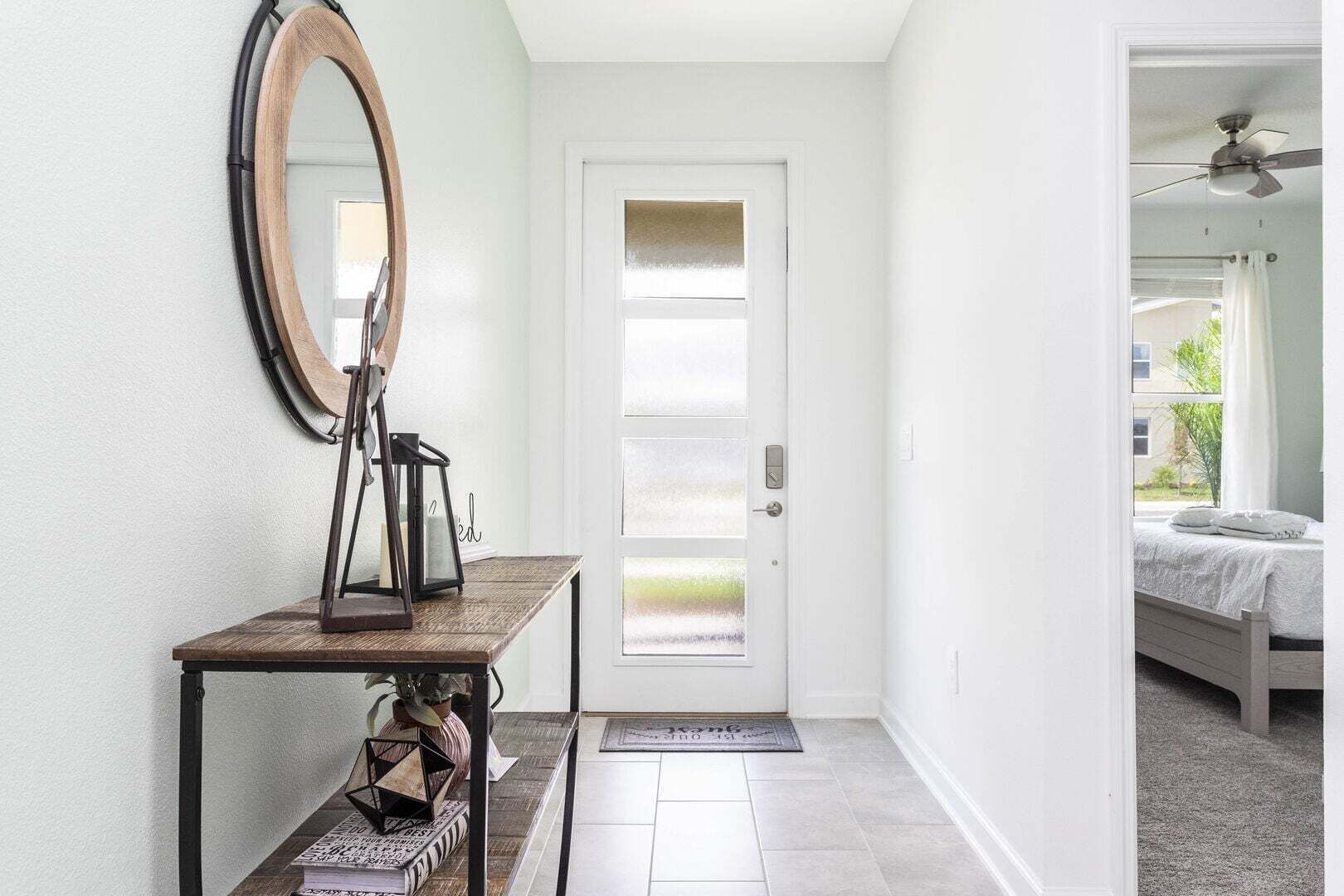 Un couloir avec une porte, un miroir, une petite table sur laquelle se trouve un vase, et un lit dans la pièce adjacente.