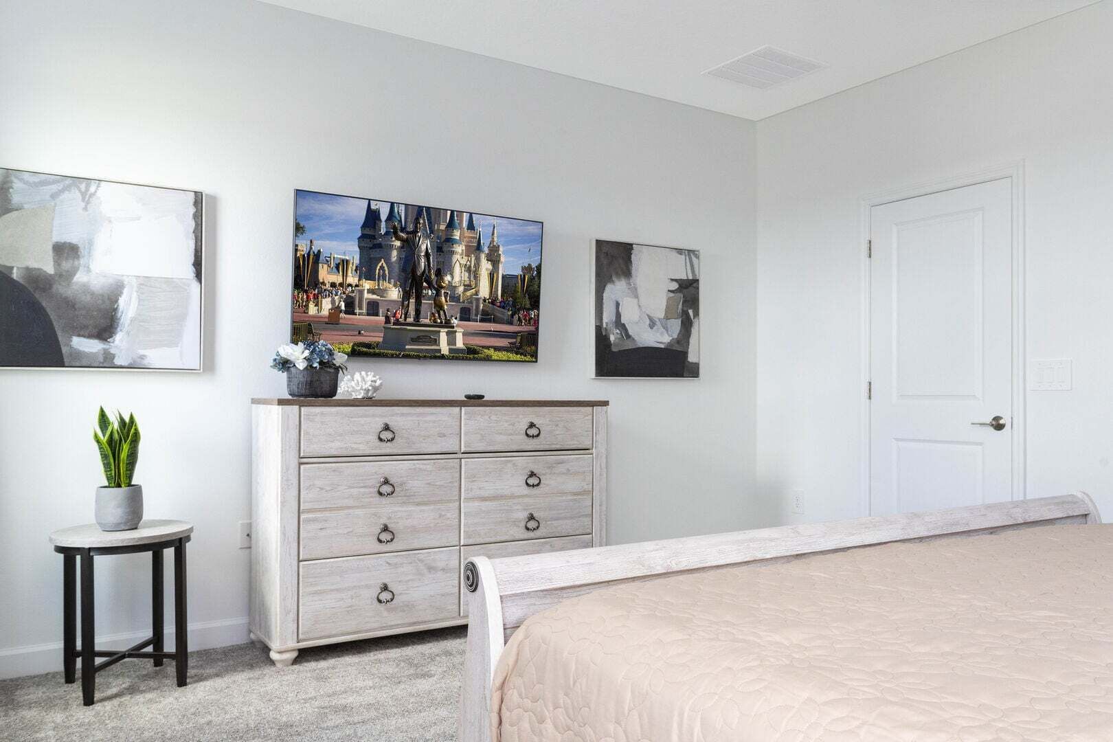 Un dormitorio con una cama, un aparador, una mesita con una planta y tres cuadros en la pared.