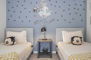 Camera da letto con due letti, ognuno con un cuscino di Topolino, un comodino con una lampada e una parete decorata a tema Topolino.