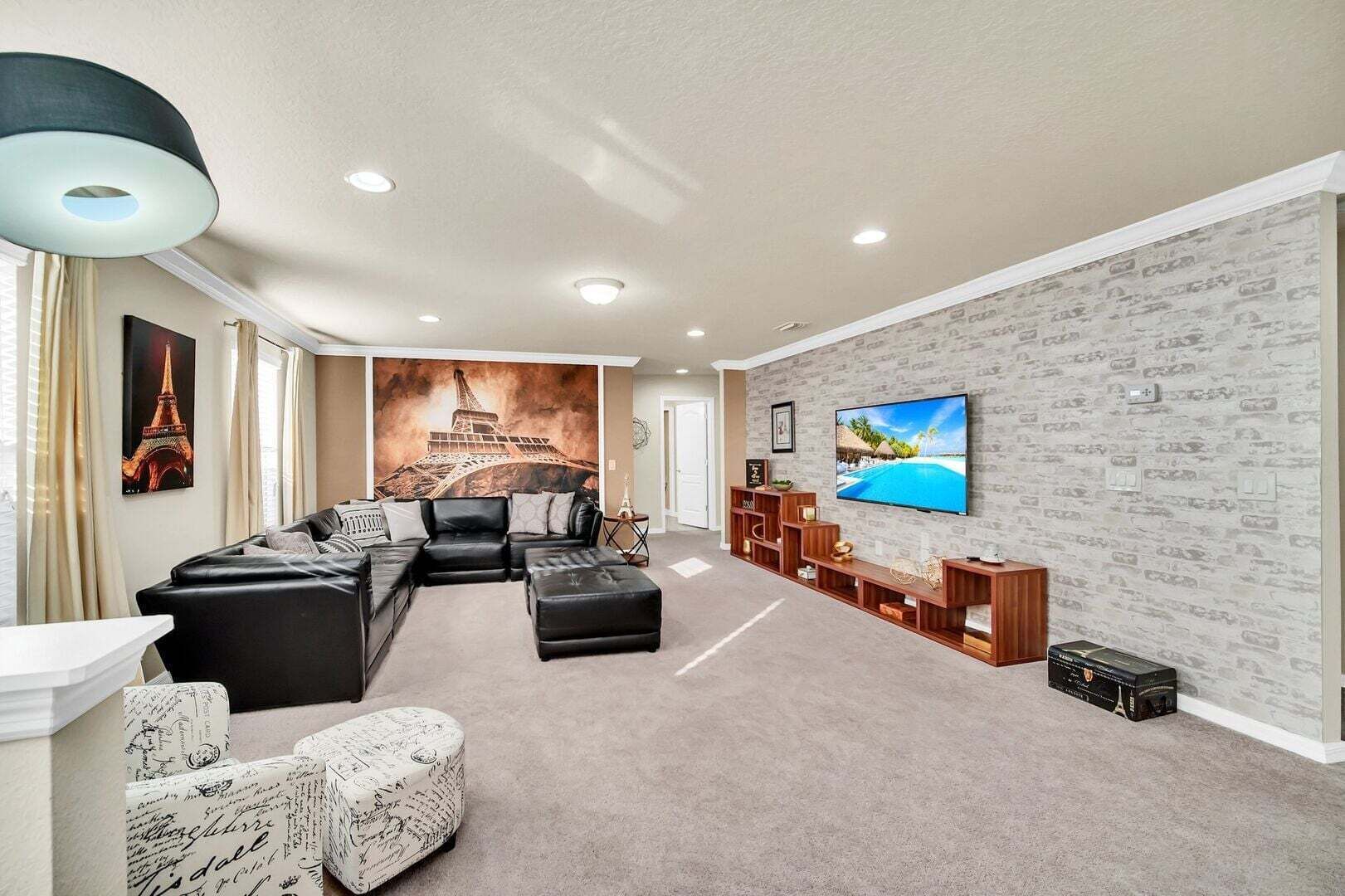 A living room with a sectional sofa, ottoman, and a flat-screen TV on a brick-patterned wall.