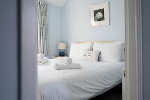 A neatly made bed with white linens and a pillow with text, a bedside lamp, and a framed picture on the wall. - The Little Blue House, Southwold (Southwold)