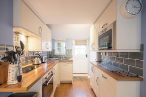 A compact kitchen with white cabinets, a microwave, and a clock on the wall. - The Little Blue House, Southwold (Southwold)