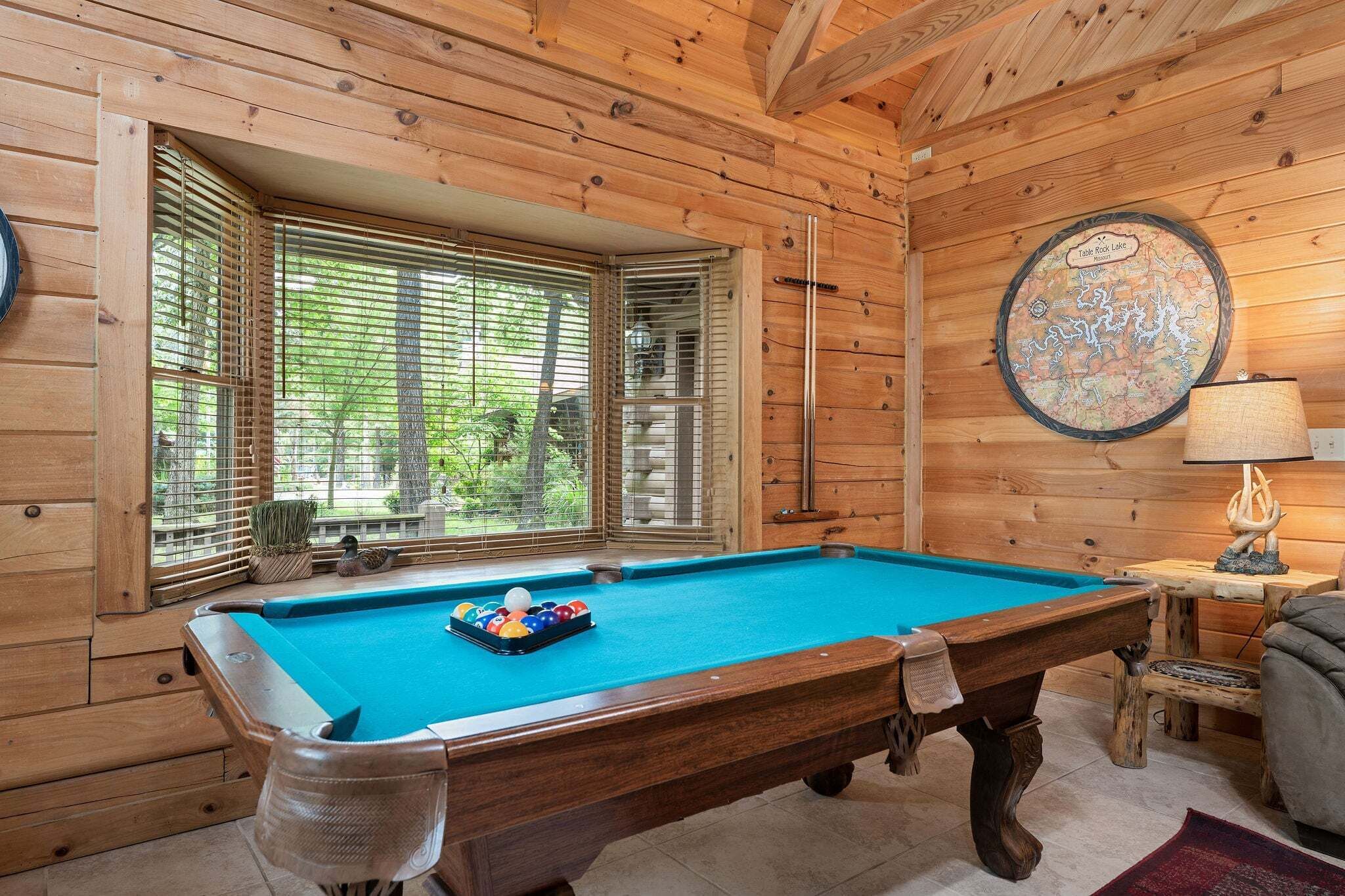 Une table de billard dans une pièce en bois, avec une carte accrochée au mur et une fenêtre équipée de stores.