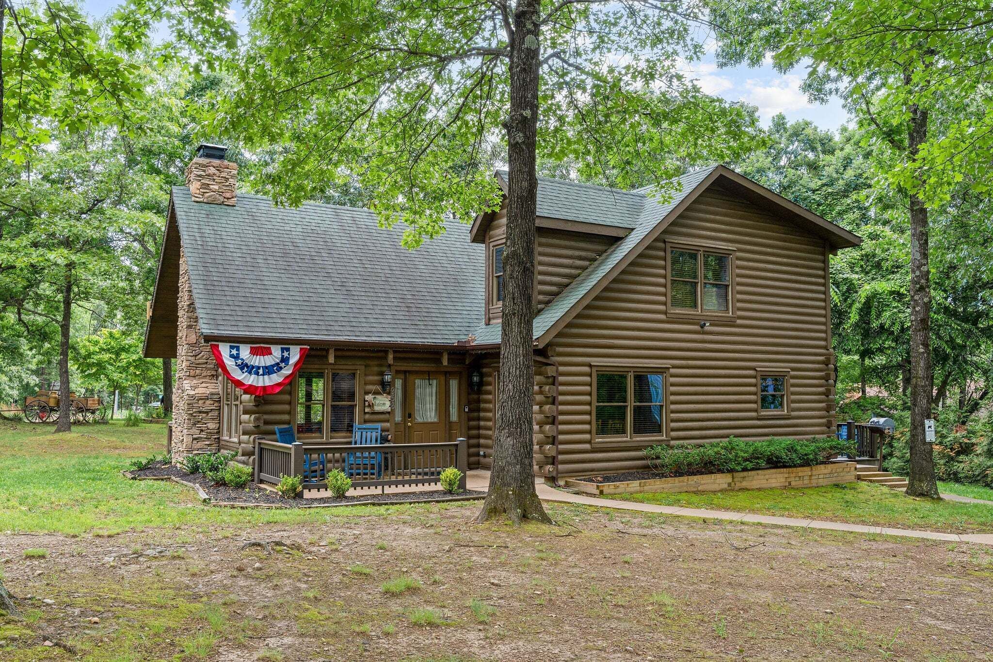 A log cabin with a stone chimney, a porch with blue chairs, and a flag.