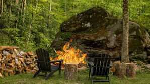 Två Adirondack-stolar, en eldstad och en hög med ved i ett skogsområde.