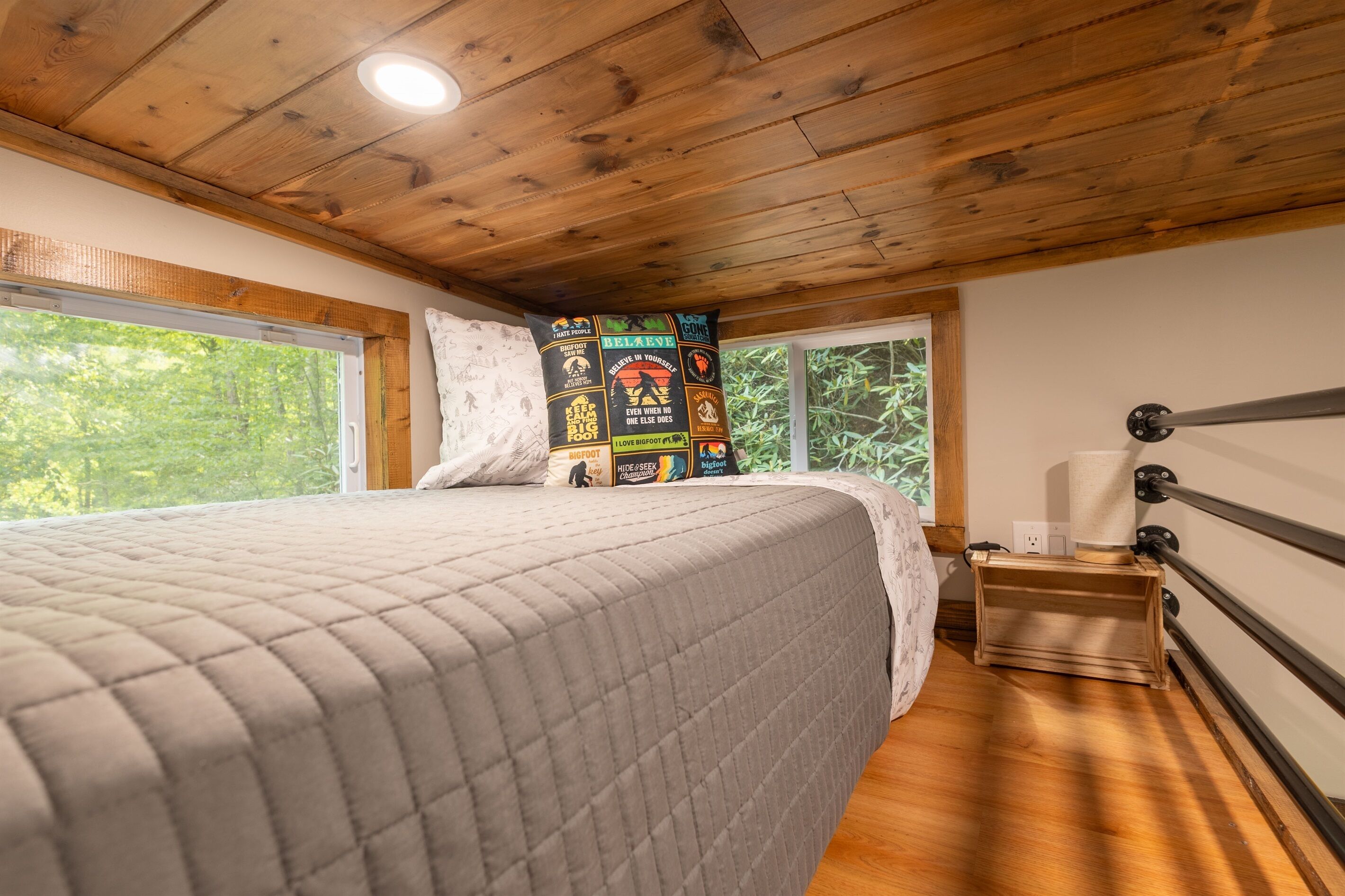 Un dormitorio con techo de madera, una cama con colcha acolchada, una ventana con vistas a árboles y una pared con parches de colores variados.