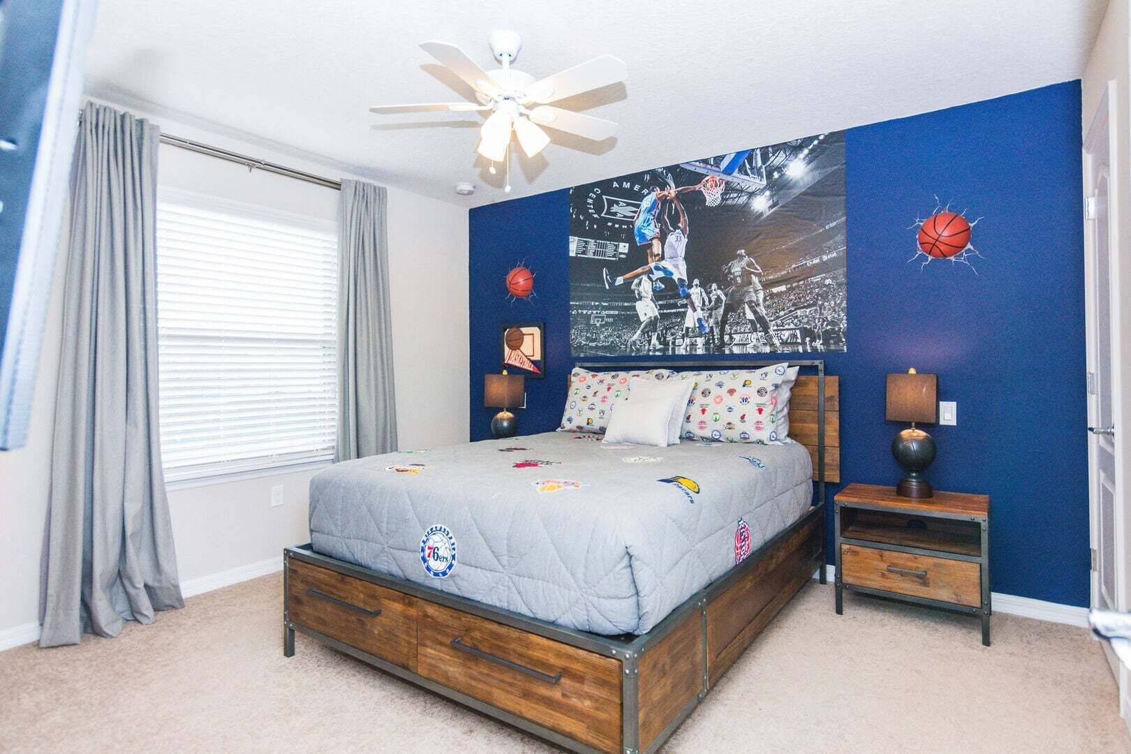 A bedroom with a bed, a ceiling fan, a basketball-themed wall, a nightstand, and a window with curtains.