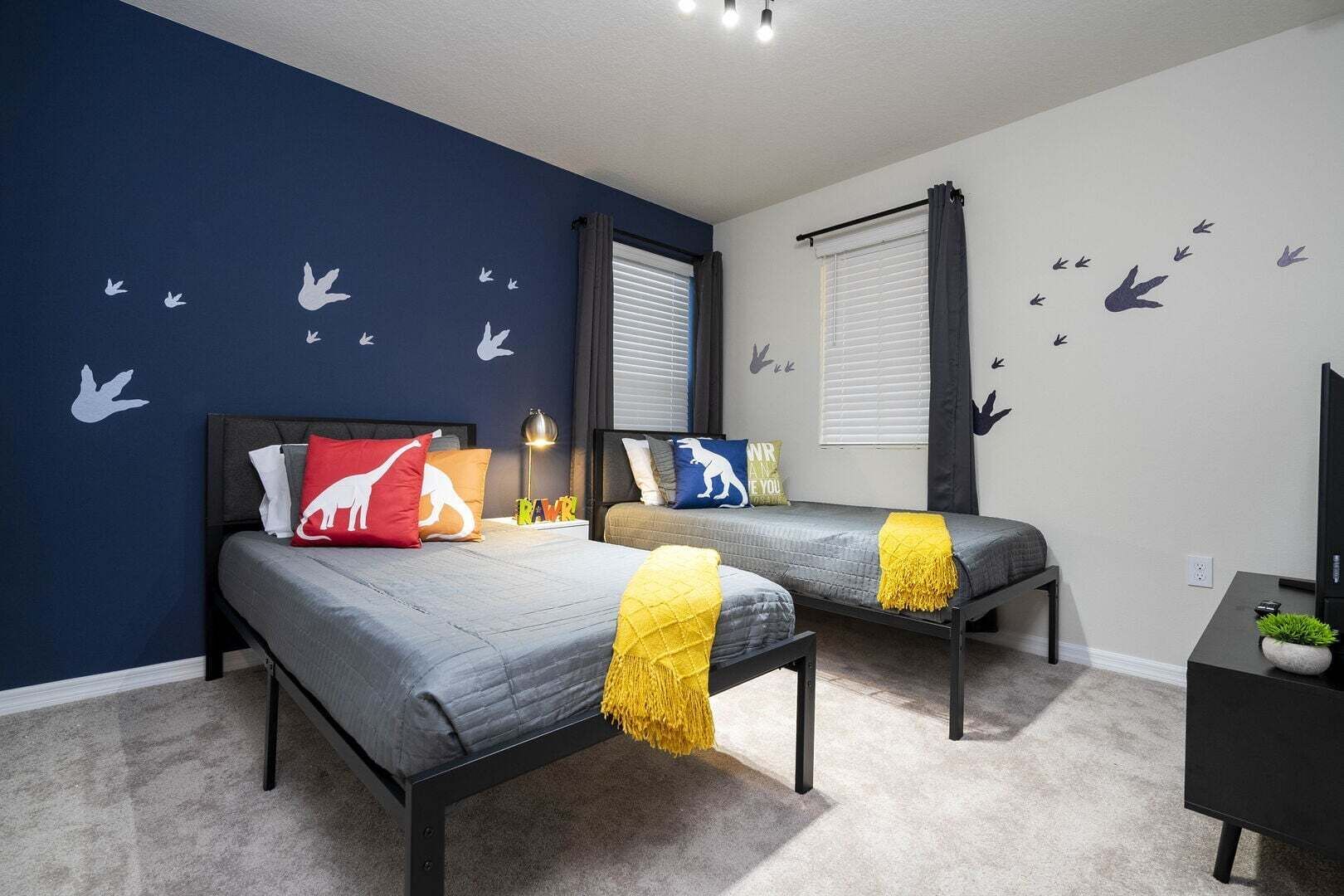 Un dormitorio con dos camas, una mesita de noche, una lámpara, un mueble para televisión y una ventana con persianas.