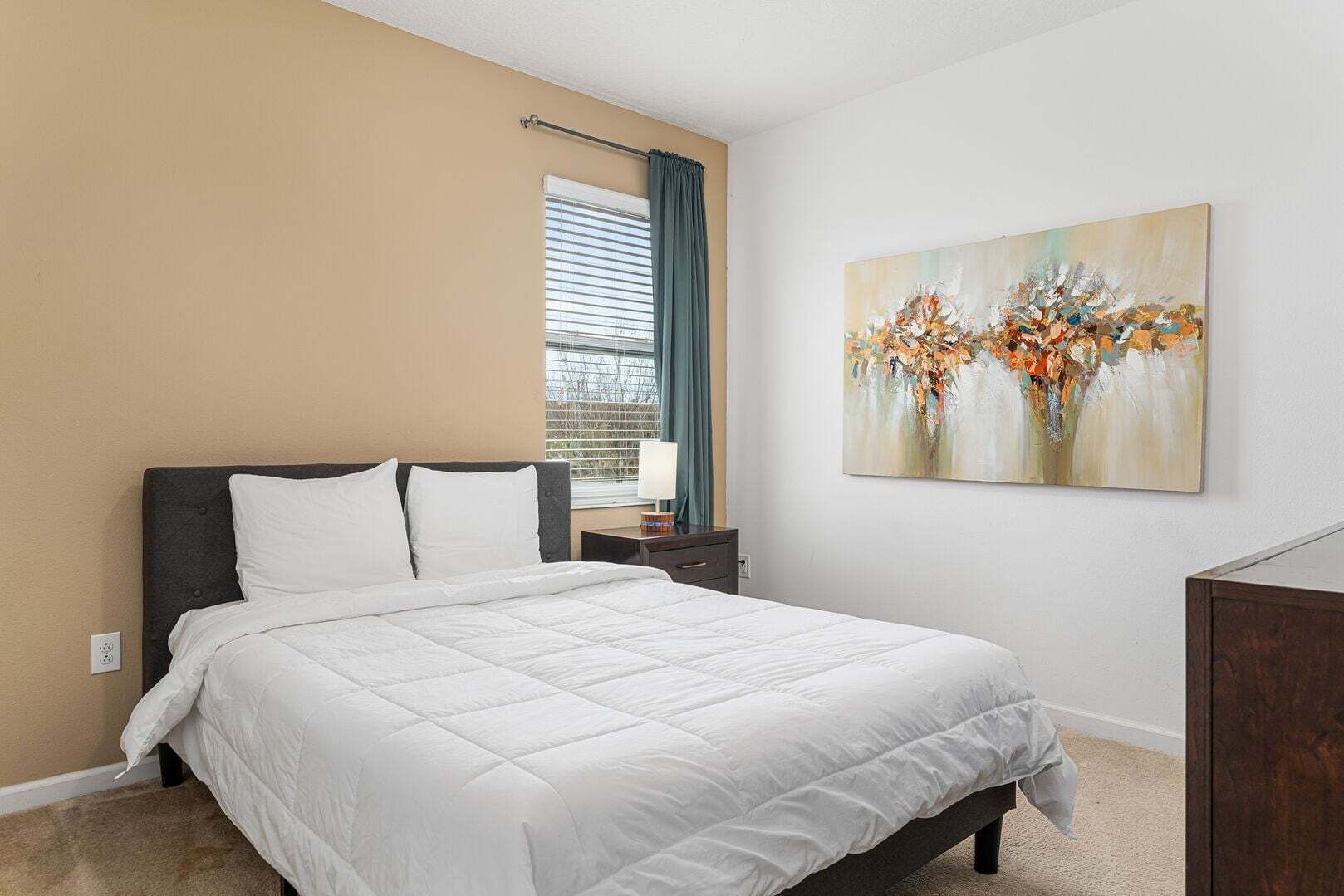 Een slaapkamer met een bed, een nachtkastje, een raam met jaloezieën en een schilderij aan de muur.