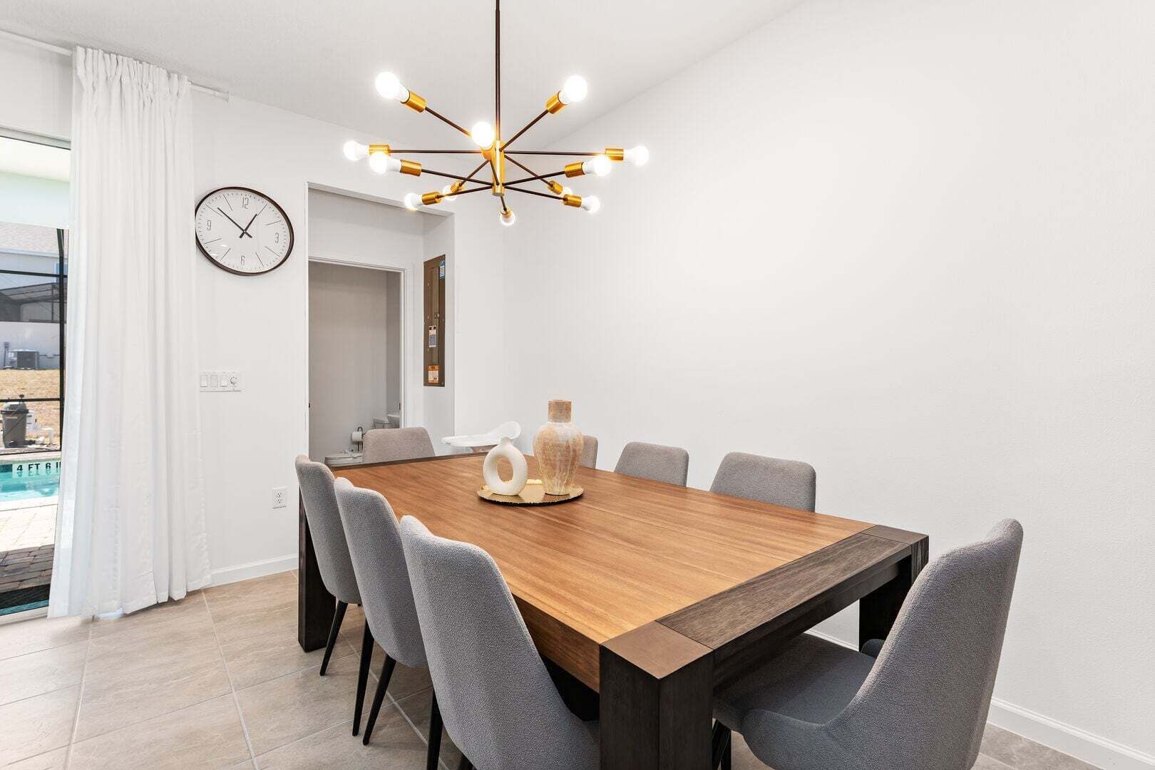 Una sala da pranzo moderna con un tavolo in legno, sedie grigie, un lampadario particolare e un orologio appeso al muro.