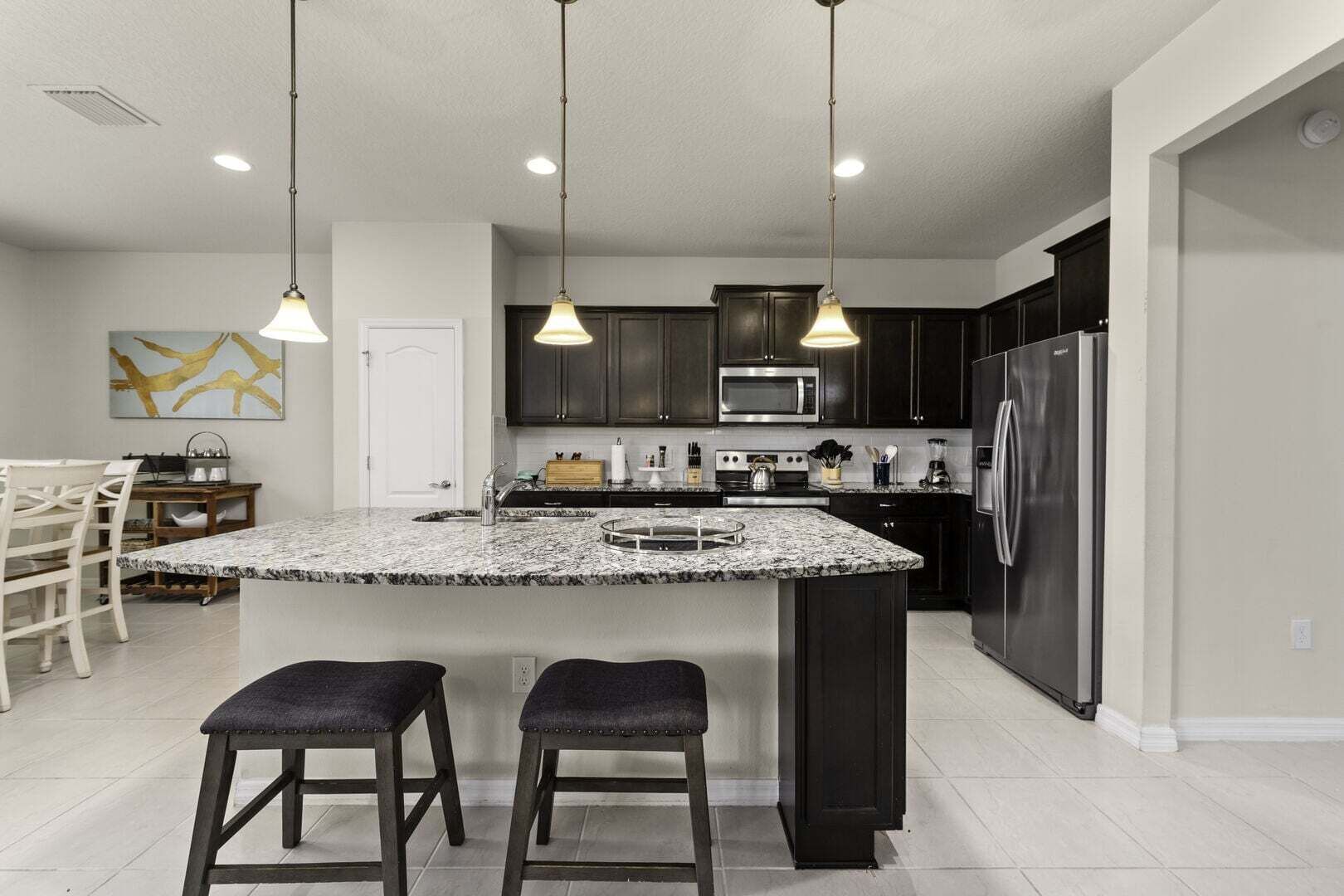 A modern kitchen with dark cabinets, a stainless steel refrigerator, a microwave, and a granite island with bar stools.