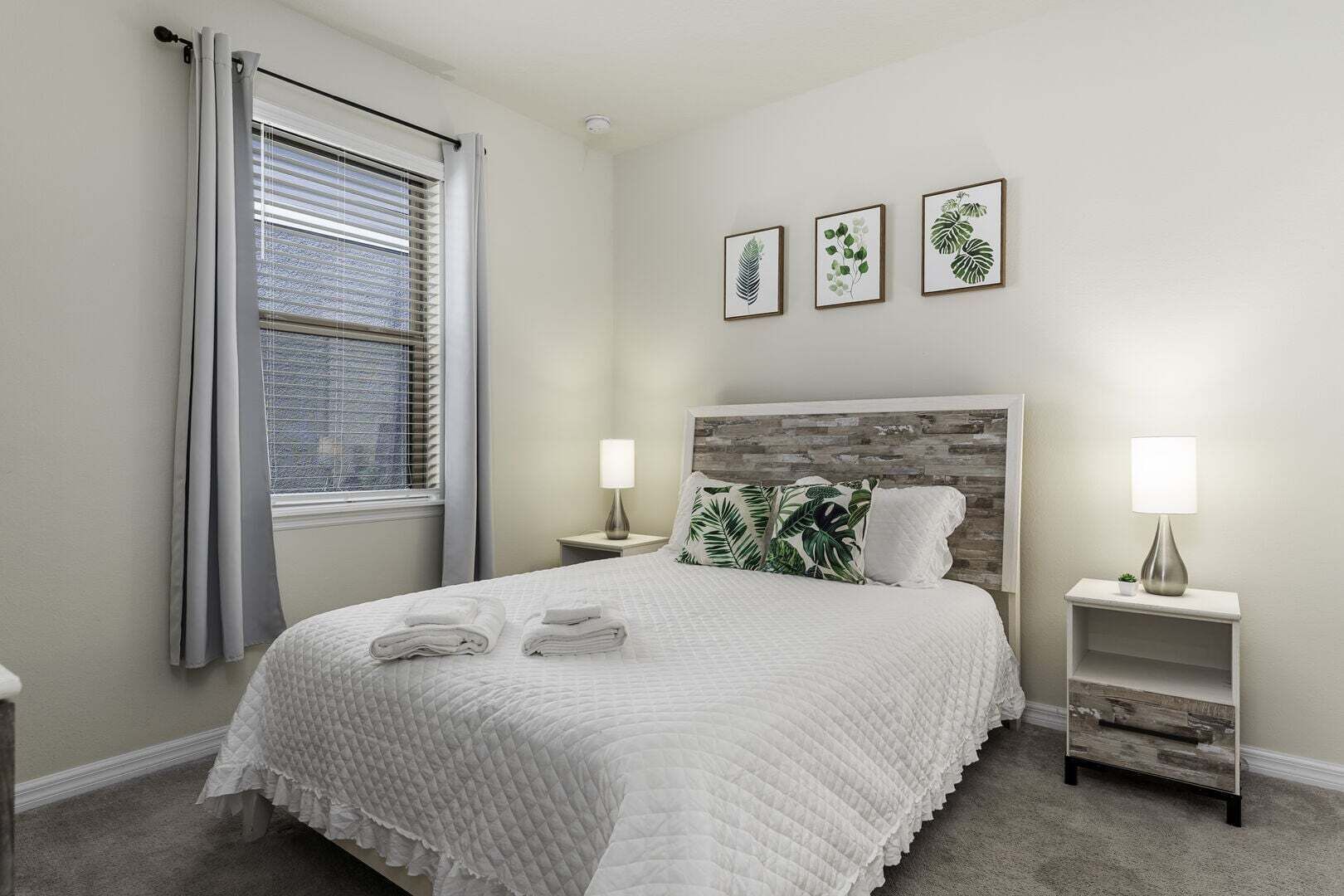 A bedroom with a bed, two bedside tables with lamps, a window with curtains, and three framed botanical prints on the wall.