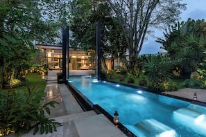 Piscina moderna con cerramiento de vidrio, rodeada de exuberante vegetación y un camino bien iluminado que conduce a un edificio.