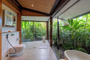 Un baño moderno con inodoro, bidé y bañera, rodeado de abundante follaje.
