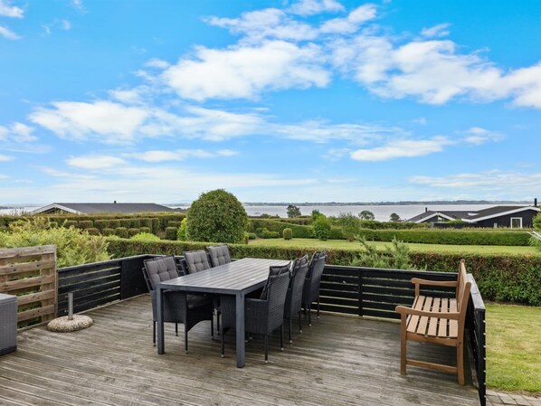 Une terrasse avec une table et des chaises, entourée d’une clôture en bois, d’où l’on peut admirer un plan d’eau et des bâtiments situés au loin.
