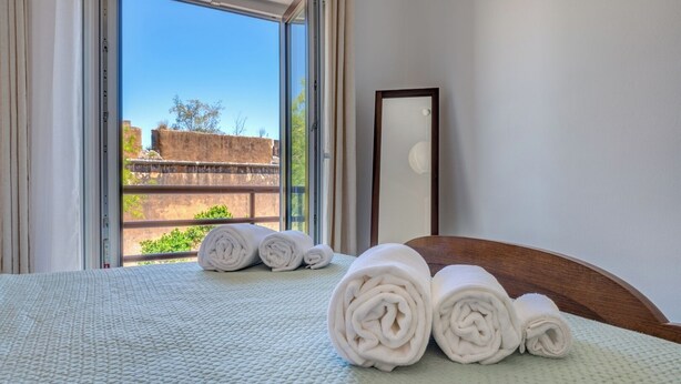 Quarto de hotel com cama, janela com vista para um edifício e vegetação, e um espelho na parede.