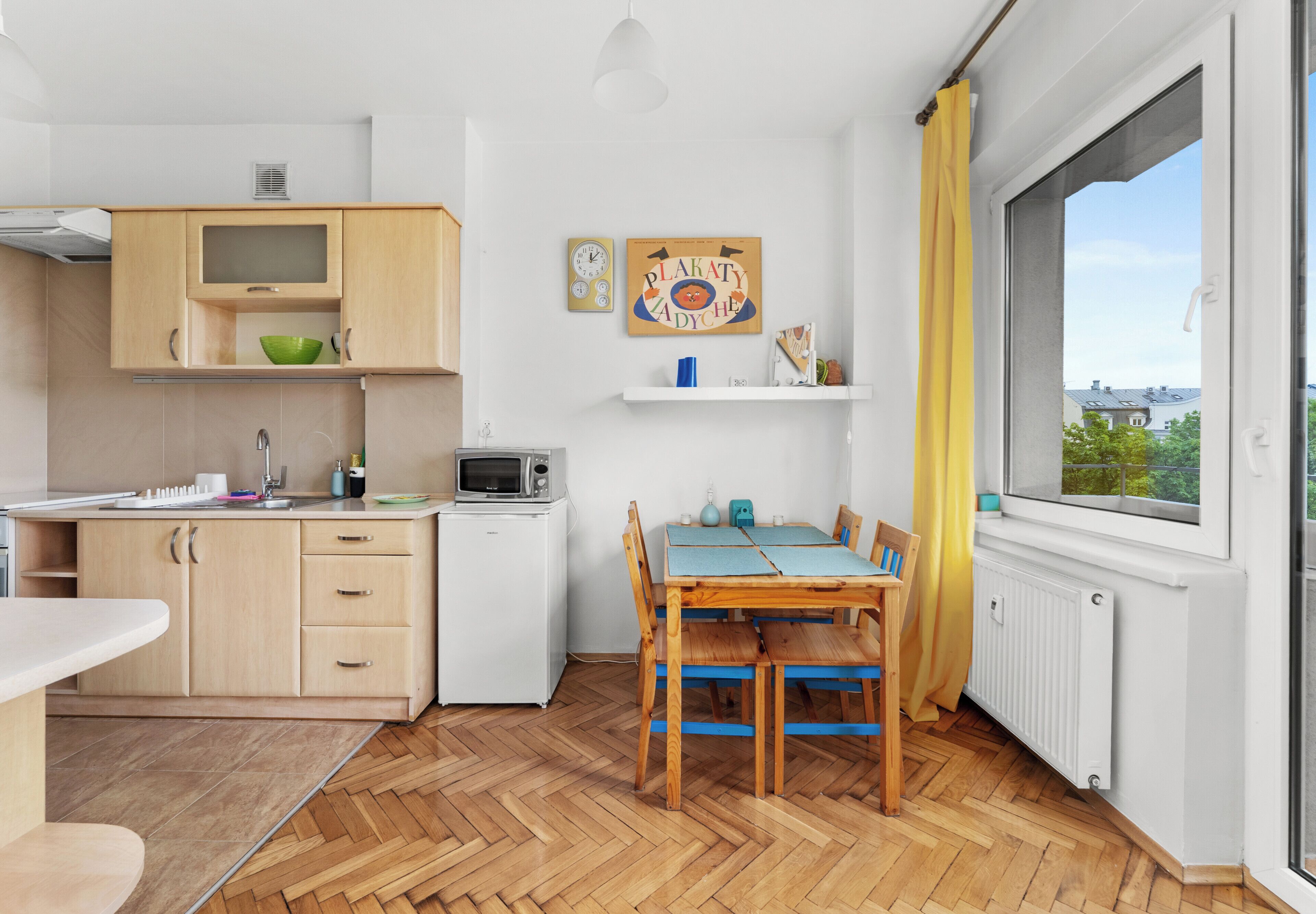 Een kleine keuken met houten kastjes, een magnetron, een kleine koelkast, een eettafel met stoelen en een raam met uitzicht.