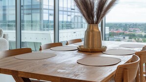 A wooden dining table with placemats and a vase with dried plants, set against a large window with a city view.