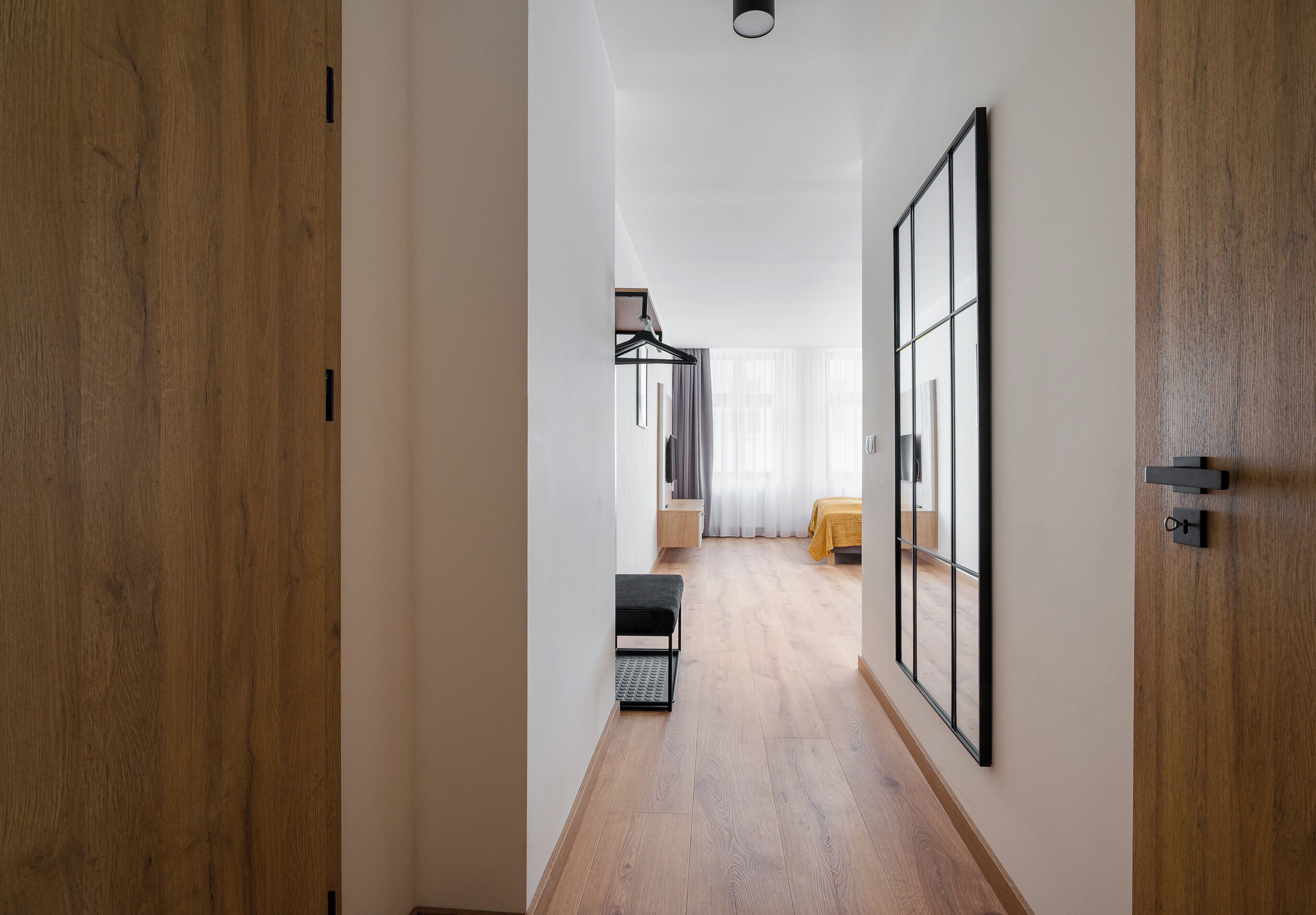 A hallway with wooden flooring, a bench, a bed with a yellow bedspread, and a large mirror on the wall.