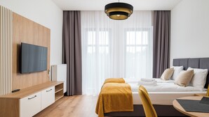 A modern hotel room with a bed, a desk, a TV, and a window with curtains. - Crocus Boutique Apartment #2 (Oświęcim)