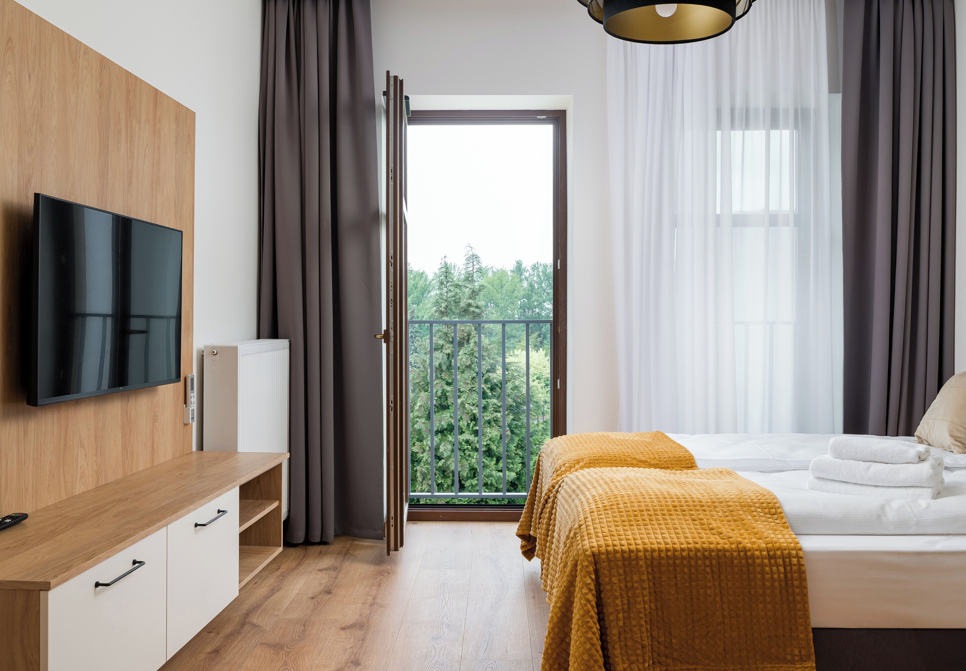 Una moderna camera d'albergo con una scrivania in legno, una televisione a schermo piatto, un letto con coperta gialla e un balcone con vista sugli alberi.