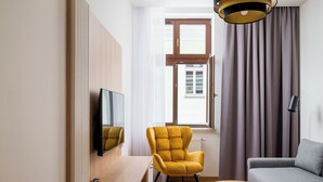 Un salon moderne avec un canapé, un fauteuil jaune, une petite table en bois, un téléviseur fixé au mur et une fenêtre avec des rideaux.