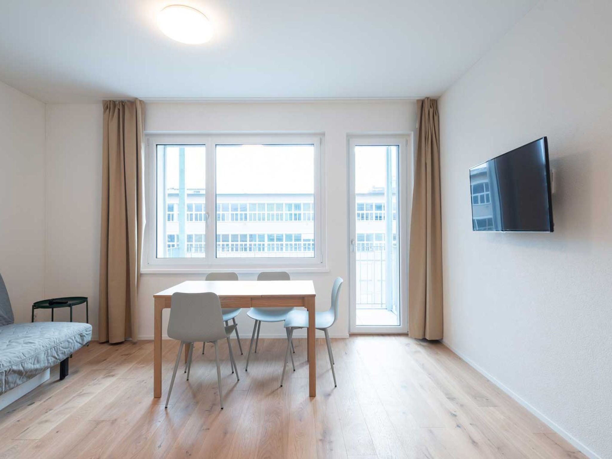A modern living room with a dining table, chairs, a sofa, a TV mounted on the wall, and large windows with curtains.