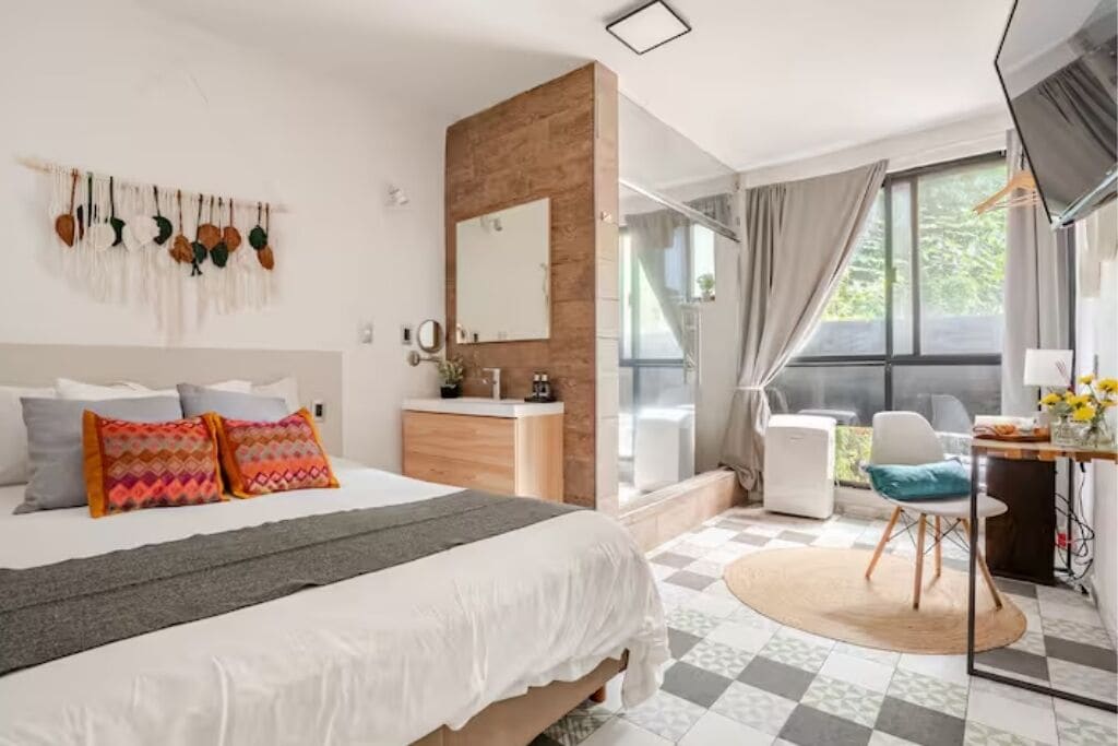 A bedroom with a bed, bedside table, mirror, and a view of the outdoors.