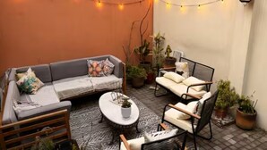 An outdoor seating area with a sofa, chairs, and a coffee table, surrounded by potted plants and string lights.