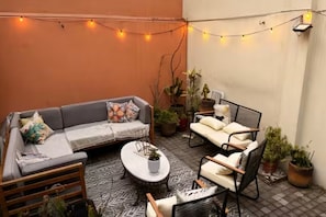 An outdoor seating area with a sofa, chairs, and a coffee table, surrounded by potted plants and string lights.