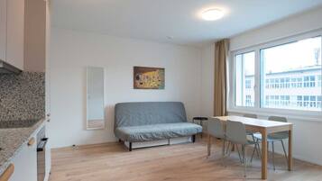 A modern, well-lit room with a grey sofa, a dining table with chairs, and a large window offering a view of buildings.