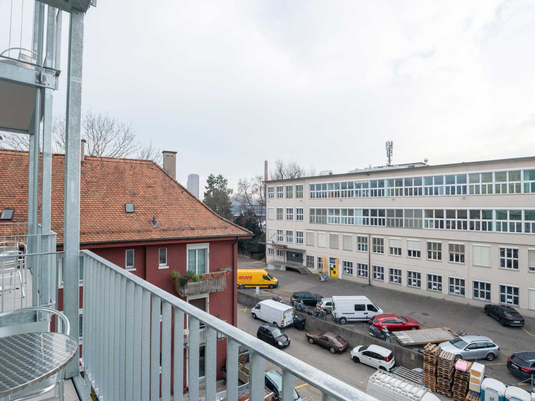 A multi-story building with a flat roof, a red building adjacent to it, and a parking lot with various vehicles.