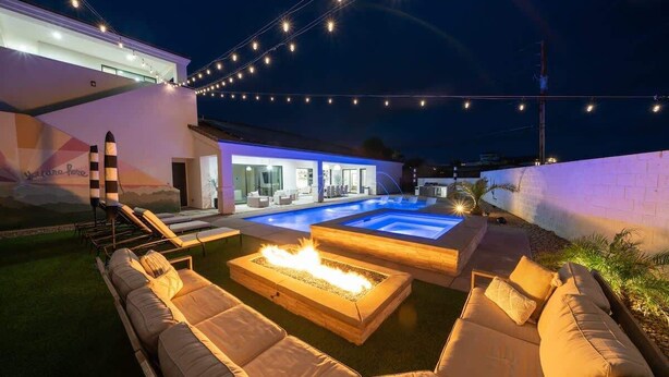 Un área de piscina moderna al aire libre con sillones de descanso, una fogata y luces colgantes.