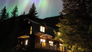 A wooden cabin with a balcony, surrounded by trees, under a night sky with aurora borealis. - Secluded North Cascades Retreat | Private Trail + Fire Pit (Marblemount)