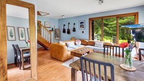 A spacious living area with a wooden staircase, a sofa, a dining table, and large windows offering a view of greenery. - Secluded North Cascades Retreat | Private Trail + Fire Pit (Marblemount)