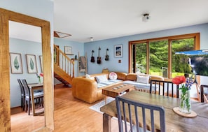 Un vaste espace de vie agrémenté d’un escalier en bois, d’un canapé, d’une table à manger et de grandes fenêtres offrant une vue sur la verdure.