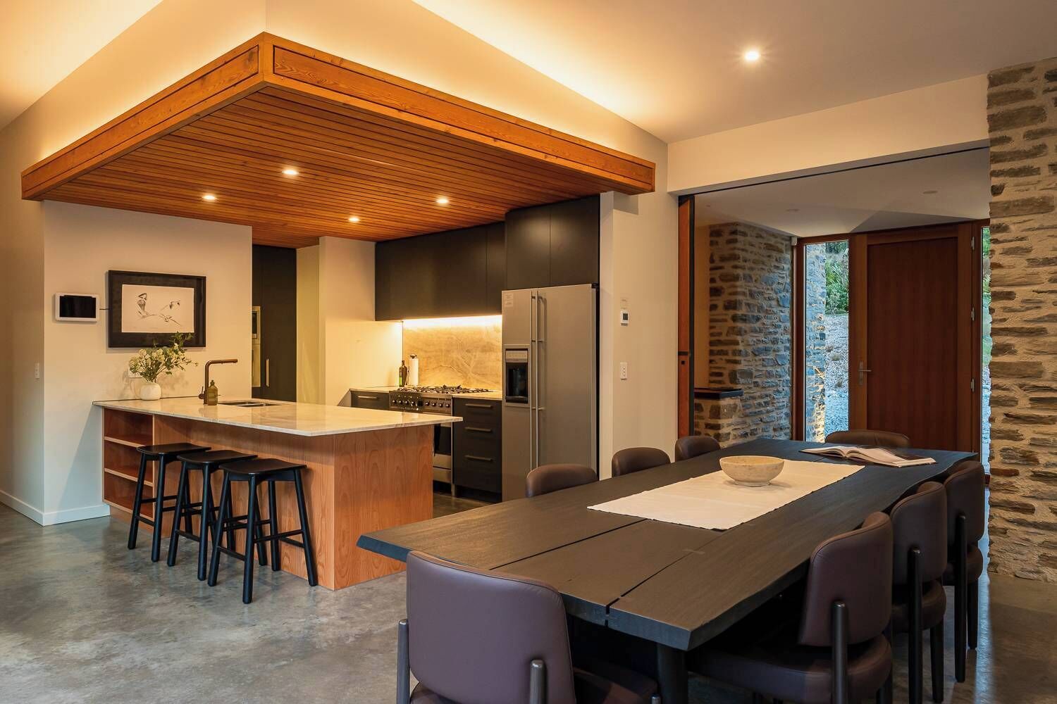 Une cuisine moderne dotée d’un îlot central, d’un coin repas avec une longue table et d’un mur en pierre.