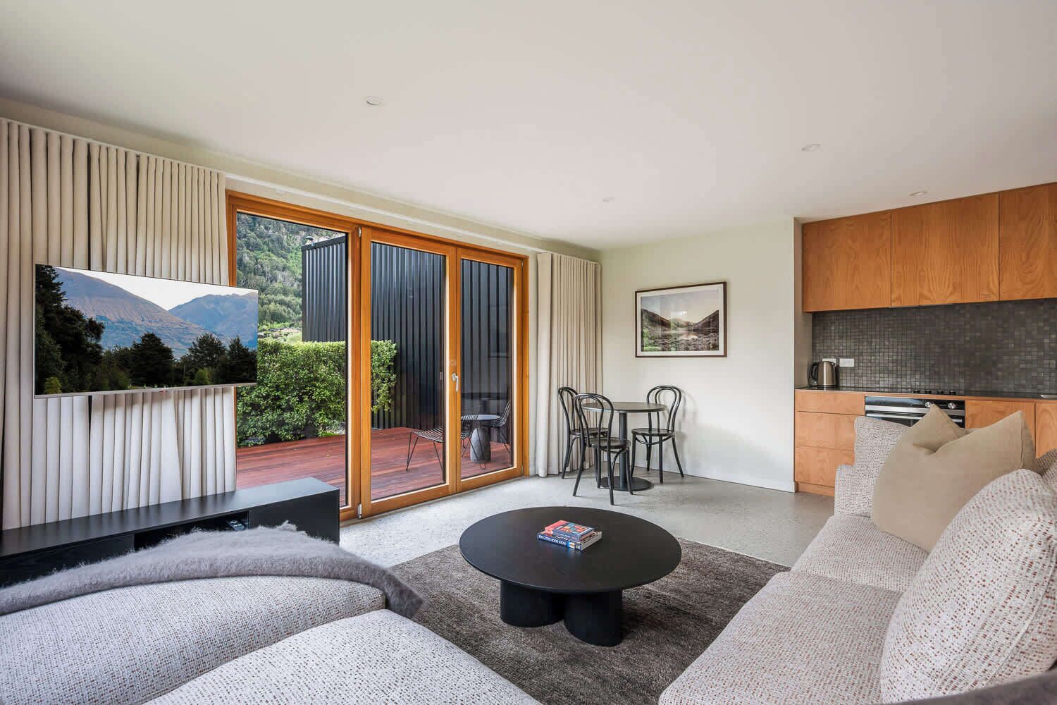 A modern living room with a sofa, coffee table, and a TV. There is a sliding glass door leading to a balcony with outdoor furniture.