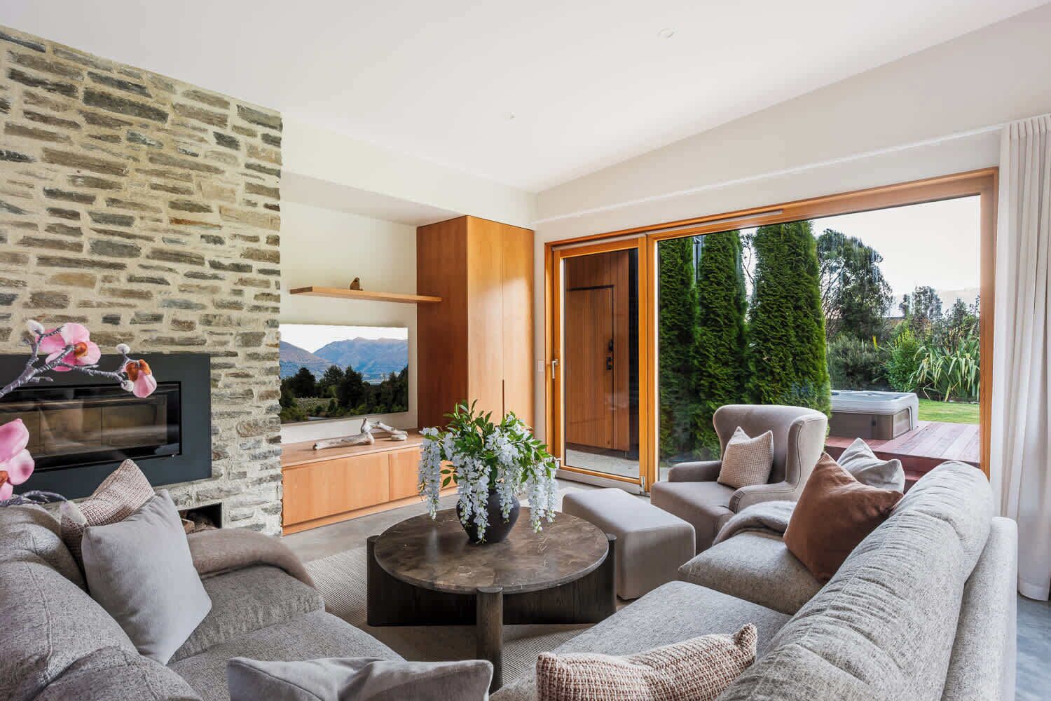 Un salon moderne avec un mur en pierre, un téléviseur à écran plat, un meuble en bois, un ensemble de canapés et une grande porte-fenêtre coulissante donnant sur un jardin.