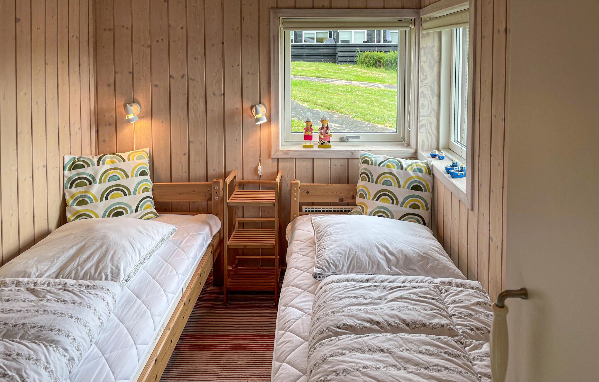 A small, cozy room with two single beds, a wooden bench, a window with a view of a garden, and a shelf with toys.