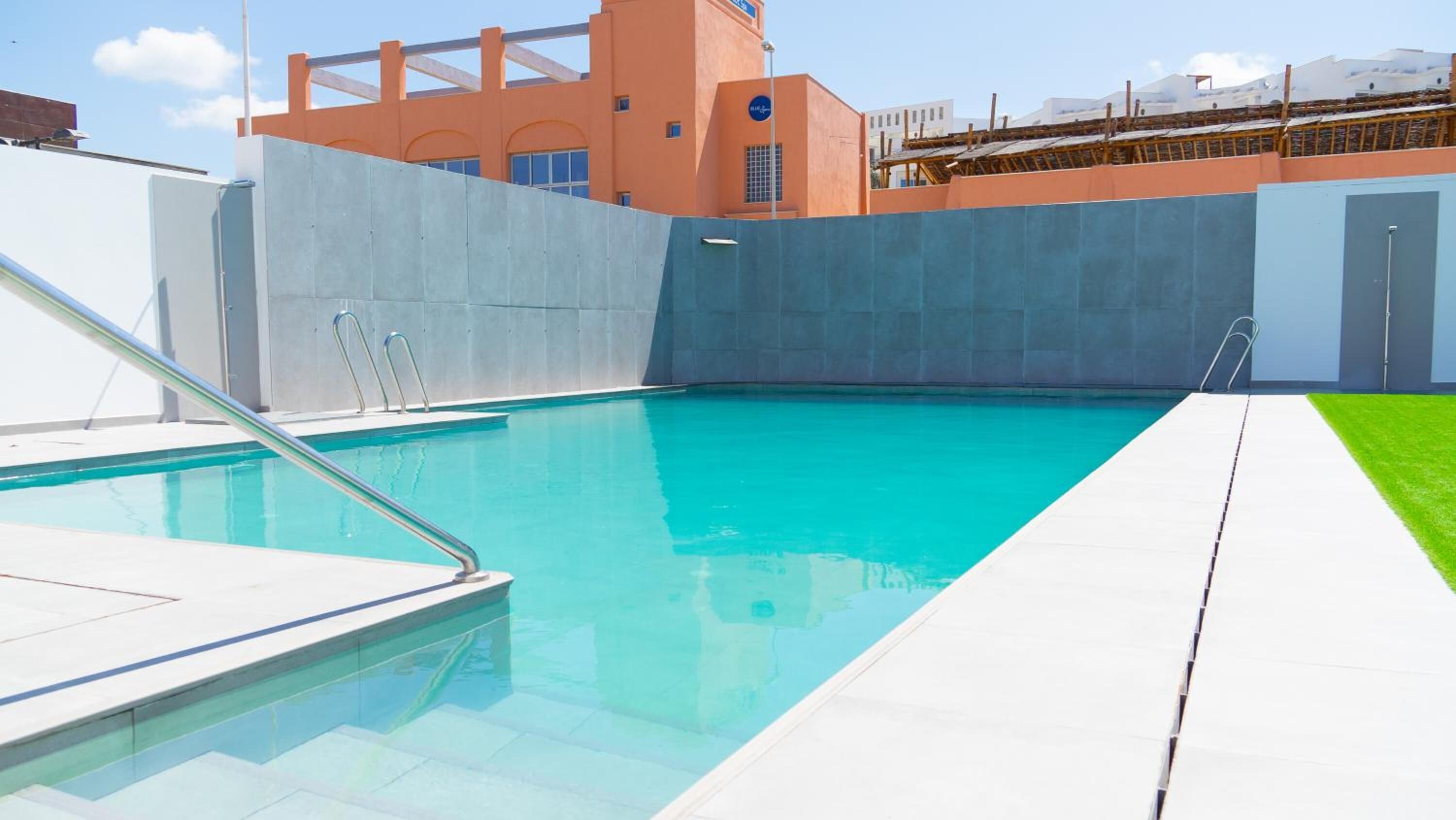 Una piscina amb un trampolí, una torre de socors i un edifici al fons amb un disseny arquitectònic singular.