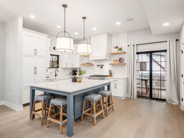 A modern kitchen with white cabinets, a central island with bar stools, pendant lighting, and a view to a balcony. - Chateau in the Village - Luxury New Build 3BD/5BA (Columbus)