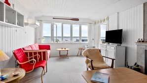 Ein Zimmer mit einem roten Sofa, einem Holzesstisch, einem flauschigen Sessel, einem Fernseher und großen Fenstern mit Aussicht.