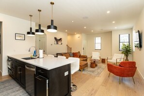 A modern kitchen with a central island, a dining area with a sofa and chairs, a living room with a coffee table and chairs, and a large window with blinds.