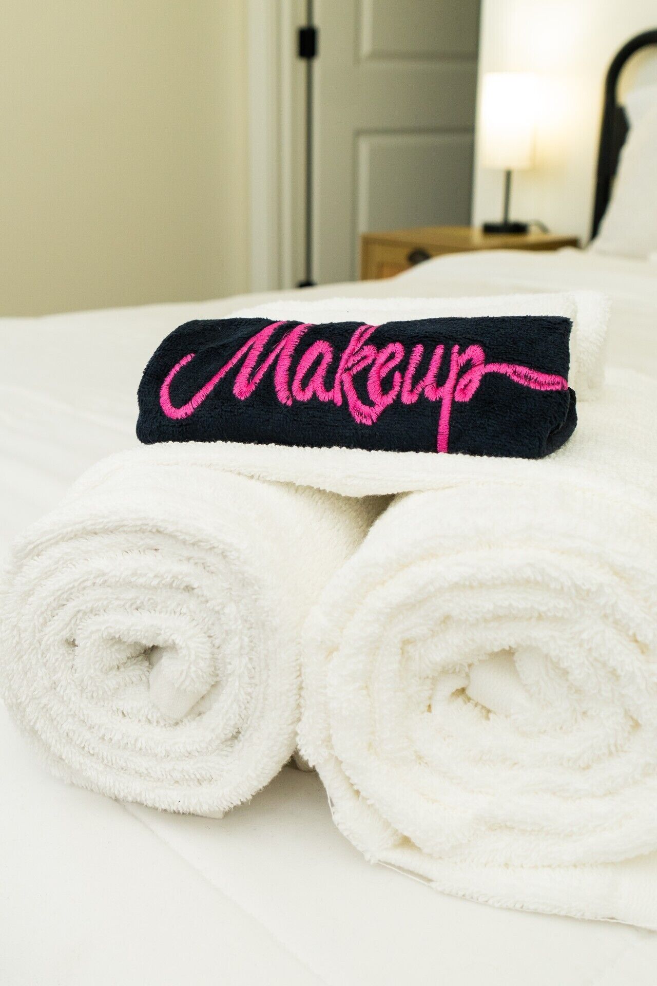 A neatly made bed with white linens and a black towel with 'Makeup' embroidered in pink.